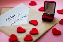 Beautiful Wedding Ring With Precious Stone In A Red Gift Box, Carved Hearts And Envelope With A Note "Will You Marry Me?" On Pink Background. Marriage Proposal Concept.