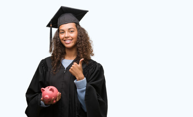 Wall Mural - Young hispanic woman wearing graduation uniform holding piggy bank happy with big smile doing ok sign, thumb up with fingers, excellent sign