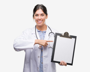 Sticker - Young hispanic doctor woman holding clipboard very happy pointing with hand and finger