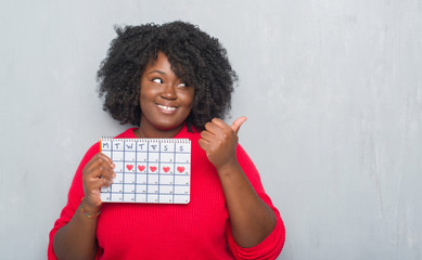 Wall Mural - Young african american woman over grey grunge wall holding menstruation calendar pointing and showing with thumb up to the side with happy face smiling