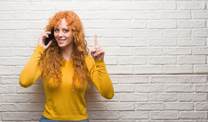 Wall Mural - Young redhead woman standing over brick wall talking on the phone surprised with an idea or question pointing finger with happy face, number one