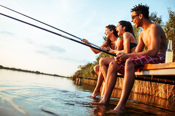 Group of friends sitting on pier by the lake and fishing.They joying in beautiful summer sunset.