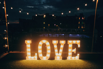 big white love letters in light bulbs for photo booth at wedding reception in night outdoors. love word lights, stylish evening decor for wedding ceremony