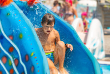 A Happy Boy On Water Slide In A Swimming Pool Having Fun During Summer Vacation In A Beautiful Aqua Park. A Boy Slithering Down The Water Slide And Making Splashes.