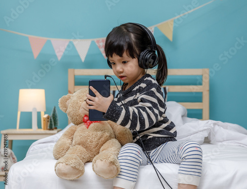 Fotografie, Obraz Cute little girl singing with smartphone with teddy bear in her bedroom, Happy a