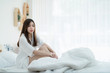 © Kanyanat - Beautiful Asian lady resting on white bed. Cute women sitting in the bed her smile thinking about something.