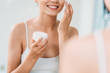 © LIGHTFIELD STUDIOS - cropped shot of smiling girl holding container and applying face cream at mirror in bathroom
