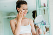 © LIGHTFIELD STUDIOS - beautiful smiling girl holding mirror and applying facial cream in bathroom