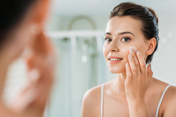 selective focus of smiling young woman applying face cream and looking at mirror in bathroom