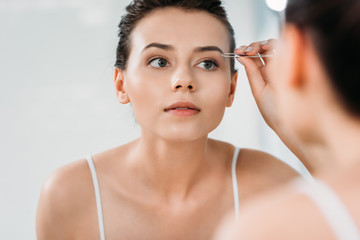 Wall Mural - selective focus of girl correcting eyebrows with tweezers and looking at mirror in bathroom