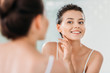 © LIGHTFIELD STUDIOS - beautiful smiling young woman touching skin and looking at mirror in bathroom