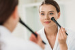 © LIGHTFIELD STUDIOS - smiling young woman looking at mirror and applying makeup in bathroom