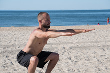 Young Athletic Man Exercising On Beach During Warm Summer Day. Squat Sit Stregth Training To Activate Core And Leg Muscles Holding Arms Straight Out.
