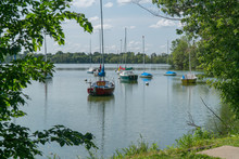 Coastal View Of Sailboats Moored In Lake Side Park On Beautiful Summer Day. View Framed Through Trees And Nature Preserve
