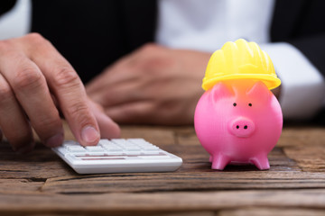 Human hand using calculator beside piggybank with hardhat