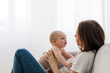 Side view of young mother sitting on sofa with baby on knees and talking on background of white wall