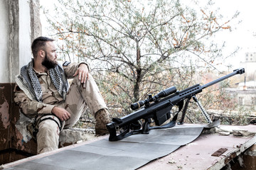 Navy SEAL sniper in battle uniform and shemagh on shoulders, sitting near large caliber sniper rifle with optics sight, on firing position in abandoned building. Urban warfare on Middle East company