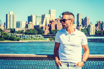 Young European Man traveling in New York, with beard, little gray hair, wearing white Polo shirt, sunglasses, standing by East River, looking around, thinking. Brooklyn buildings on far background..