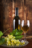 Fototapeta Panele - Table white wine in glass and black bottle on desk with grape and vine decoration. Old wooden retro background. Vintage food and drink concept. Close up, selective focus