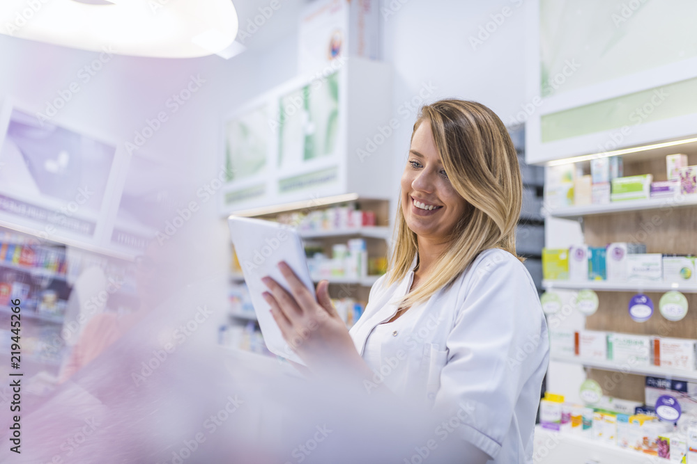 Pharmacist working with a tablet computer in the pharmacy holding it in her hand while reading information - obrazy, fototapety, plakaty 