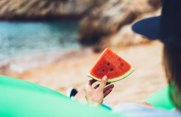 girl hipster relax on beach coast and holds in hand a slice of red fresh fruit watermelon on blue sea background, woman on seaside nature eating sweet healthy food, vacation in summer concept