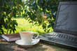 cup of coffee, a computer on a wooden table in the garden on a sunny afternoon with a copy space, a concept of working in nature freelancing