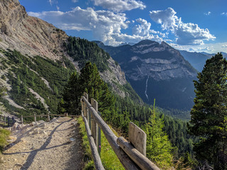 A wood railed path on a hillside of a mountain in the Alps with a view on mountains, valley and the blue cloudy sky