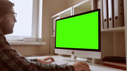 Leinwandbilder - man sitting at workplace paying game on computer in small office. businessman with beard looking on display with green screen. employee in startup company typing on keyboard use pc mouse