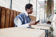 © AS Photo Family - African man in traditional clothes and glasses sitting behind laptop at outdoor caffe and looking on mobile phone.