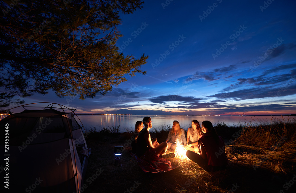 Night summer camping on sea shore. Group of five young tourists sitting on the beach around campfire near tent under beautiful blue evening sky. Tourism, friendship and beauty of nature concept. - obrazy, fototapety, plakaty 