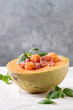 Melon and ham or prosciutto salad served in half of Cantaloupe melon, decorated by fresh basil standing on white tablecloth.