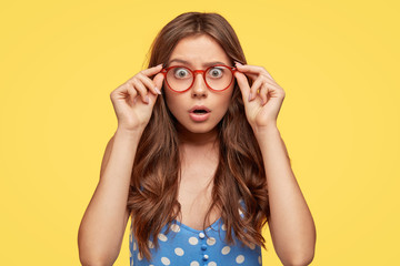 Wall Mural - Shocked attractive teenager wears glasses with red rim, polka dot blouse, stares at camera with frightened expression as hears horrified news, poses against yellow background. Omg, its dreadful!