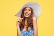© wayhome.studio  - Good looking comely merry woman wears stylish summer hat, blue polka dot clothes, holds shades, being impressed by unforgettable vacation, poses indoor against yellow wall. People, holidays concept