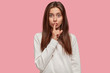 © WHstudio Leushin N - Lovely green eyed brunette female asks to keep confidential information in secret, gestures with index finger on mouth, wears loose white sweater, poses against pink background. Conspiracy concept