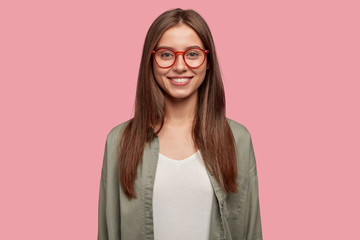 Wall Mural - Studio shot of attractive student has gleeful expression, toothy smile, being in high spirit after lectures, dressed casually, wears round spectacles stands against pink background. Lively girl indoor