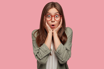 Wall Mural - Terrific young female stares with bugged eyes, keeps hands on both cheeks, recieves bad news from interlocutor, wears round spectacles, isolated over pink background. People, amazement, shock