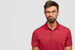 Angry person with sullen surprised expression, purses lips, raises eyebrows in bewilderment, being unshaven, stands against white background with copy space for advertisement. Facial expressions