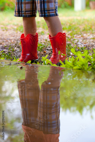 Fotografia, Obraz Legs of child wearing pair of red rubber boots with reflection in water puddle