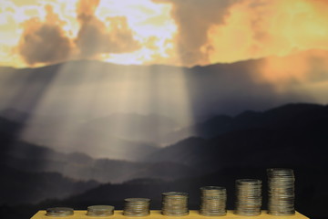 funding for environmental conservation.  pile of coins in increasing chart shape in front of dramatic god light beam from bursting cloud over mountain in the background.