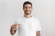 © Damir Khabirov - Daylight shot of smiling young man pointing at his blank white t-shirt with index finger, copy space for your ads, isolated on gray background