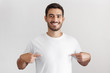 © Damir Khabirov - Smiling nice man pointing at blank white t-shirt with both index fingers, copy space for your advertising, isolated on grey background
