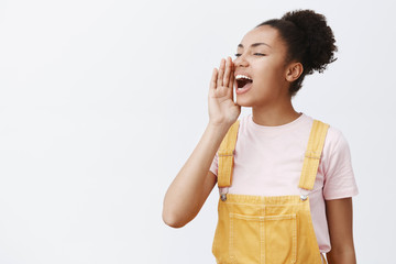 Girl shouting out loud to call friend come downstairs help her. Portrait of charming carefree urban female in yellow trendy overalls, turning left and holding palm near opened mouth, yelling