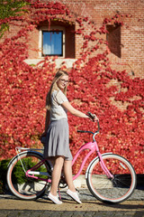 Young attractive smiling blond woman in casual clothing and glasses posing at pink lady bicycle on bright warm sunny day on background of brick wall overgrown with beautiful red ivy leaves.