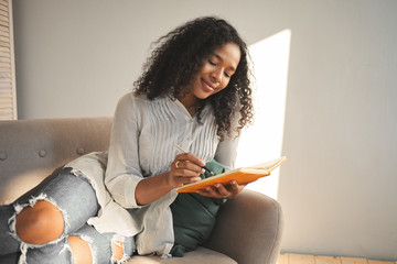 Candid shot of charming pretty girl of mixed race appearance smiling while writing down poems, being in love, sharing her thoughts and secrets with diary. People, lifestyle and leisure concept