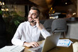 © Seventyfour - Portrait of young bearded businessman speaking by phone and smiling happily while working with laptop in cafe