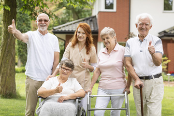 Poster - Thumbs up shown by happy female and male seniors and a caregiver outside their nursing home.