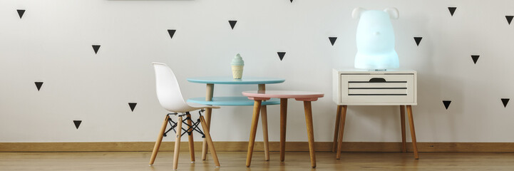 Two pastel pink and blue tables and white chair standing in real photo of bright kid room interior with animal shape lamp with cupboard