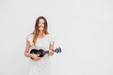 Cute young woman plays ukulele against the background of a white wall and smiles.