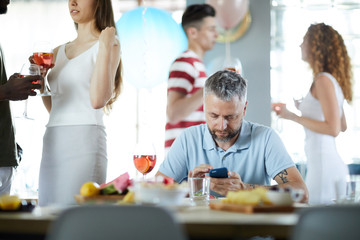 Young man with smartphone sitting by table and texting while his friends enjoying party