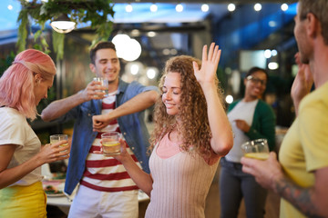 Excited young woman with drink dancing among group of friends and enjoying party